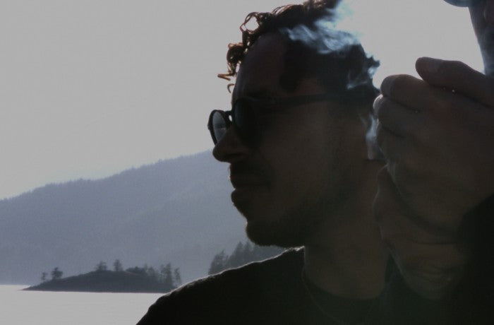 Person mit Sonnenbrille entspannt am See mit Bergkulisse und medizinischem Cannabis in der Hand.