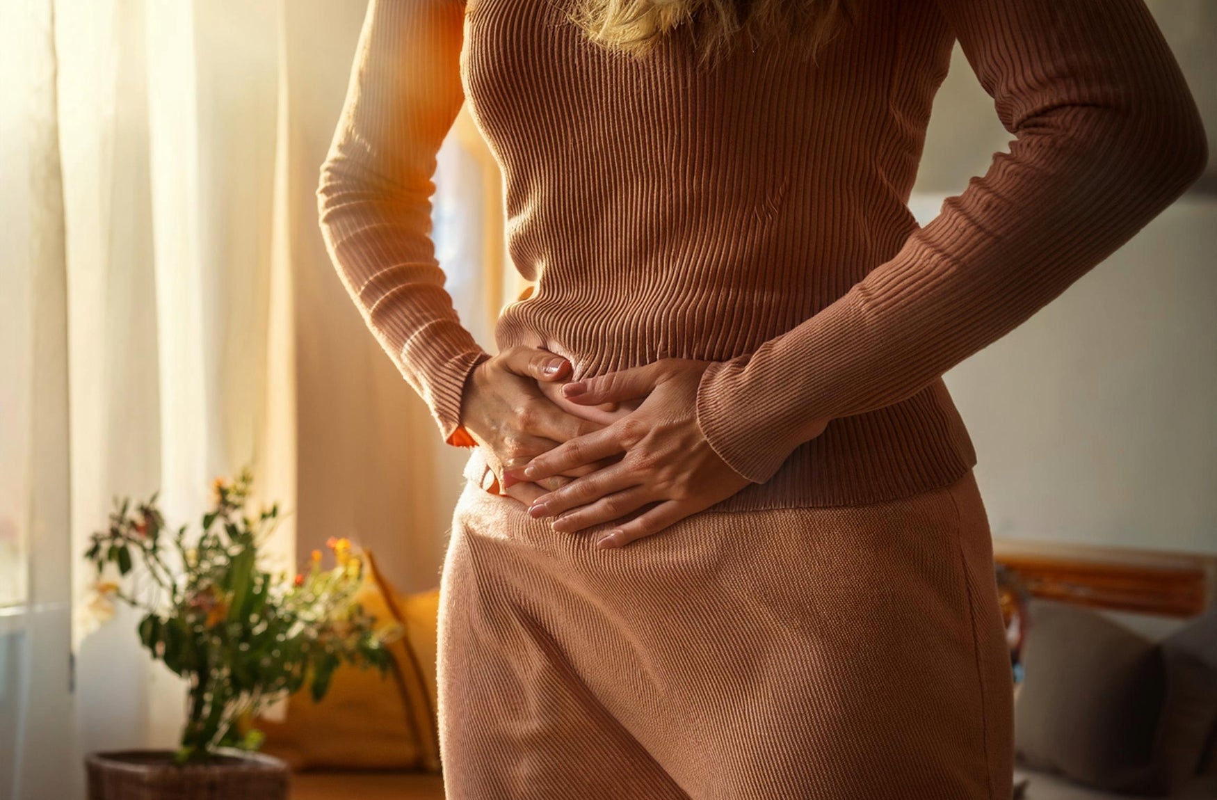 In einem Raum steht eine Frau mit Unterleibschmerzen, sie zeigt Anzeichen von Unbehagen und hält sich den Unterleib.